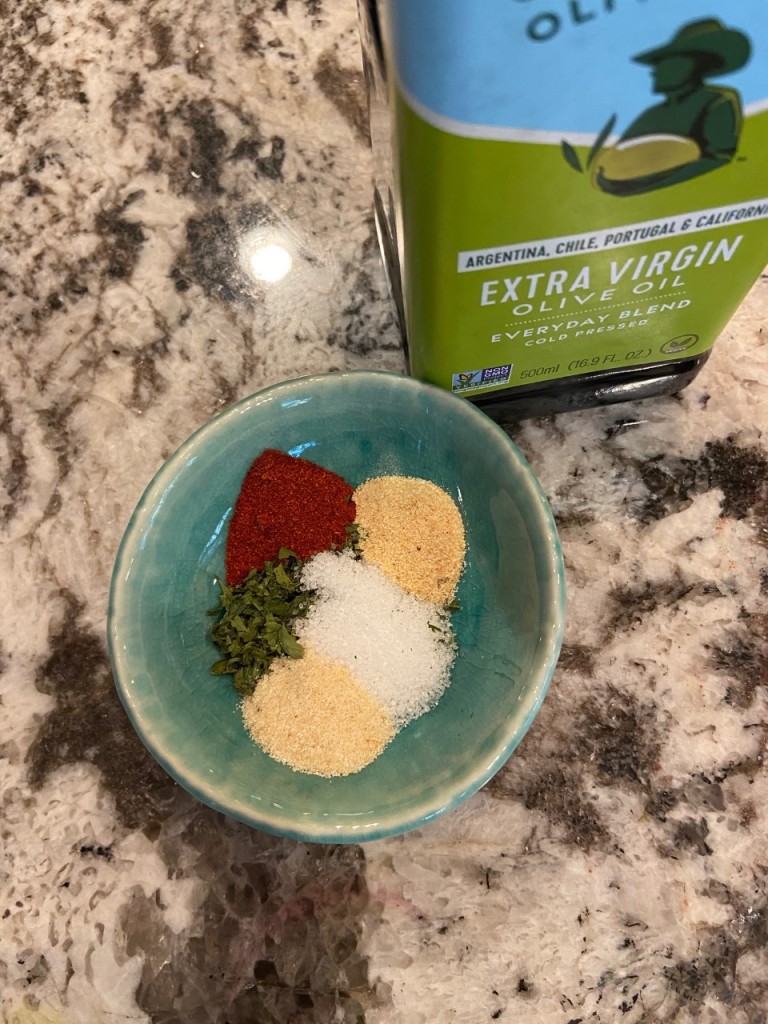 Small bowl of spices used in the recipe and a bottle of extra virgin olive oil.